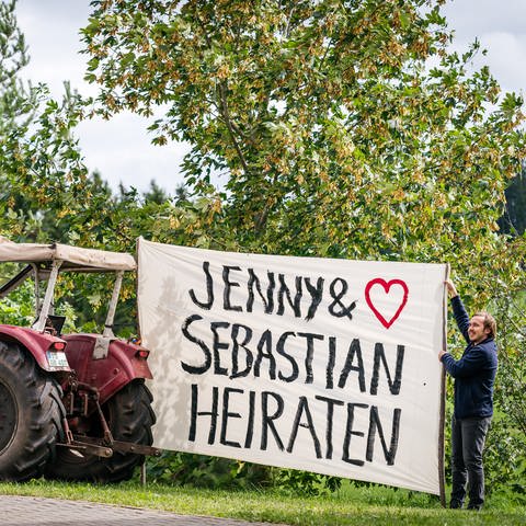 Sebastian hält ein riesiges Stück Stoff, das an einem Traktor befestigt ist. Darauf steht "Jenny und Sebastian heiraten"
