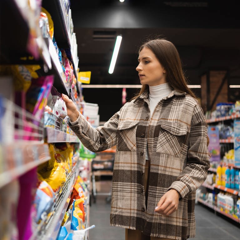 Eine Frau guckt sich Lebensmittel im Supermarkt an. Katjes, Oreo, Sanella – im Supermarkt entdecken wir immer wieder versteckte Preiserhöhungen. Die aktuellen Tricks der Hersteller.   