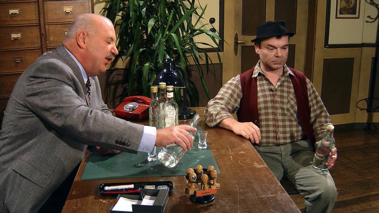 zwei Männer sitzen in einem altmodischen Büro und trinken Schnaps