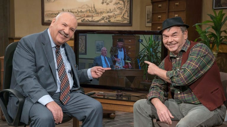 Zwei Männer sitzen vor dem Fernseher und zeigen auf einen Sketch, in dem die beiden im gleichen Ambiente, einem altmodischen Büro, mitspielen