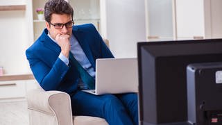 Ein Mann im Anzug mit einem Laptop auf dem Schoss schaut gebannt auf einen Fernseher