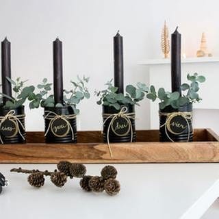 Etwas Grün, wie Moos oder Eucalyptus um die Kerzen herum stecken. Alle vier Dosen auf das Holztablett stellen und wer es gern üppiger mag, kann das Tablett mit zusätzlicher Dekoration bestücken.