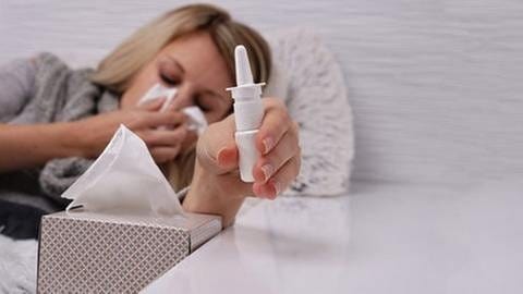Eine Frau hält sich mit geschlossenen Augen ein Taschentuch vor Nase und Mund und greift nach einem Nasenspray