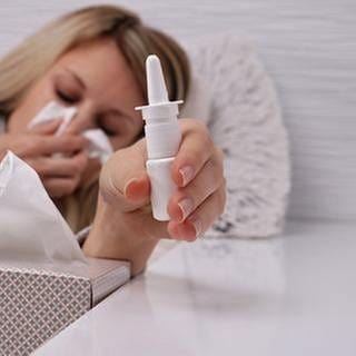 Eine Frau hält sich mit geschlossenen Augen ein Taschentuch vor Nase und Mund und greift nach einem Nasenspray