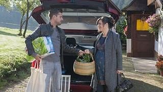 Andreas und die schwangere Eva kommen vom Einkaufen und stehen hinter dem geöffneten Kofferraum ihres Wagens