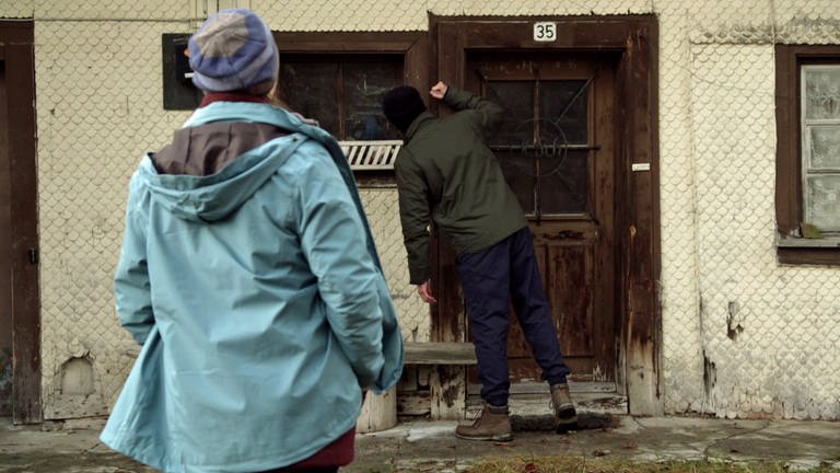 Constantin klopft an die Tür eines heruntergekommenen Bauernhofs, Sophie steht hinter ihm