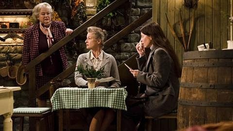 Johanna sitzt mit der weinenden Frau Rabenalt an einem Tisch im Hofladen, Leni steht hinter Johanna auf der Treppe
