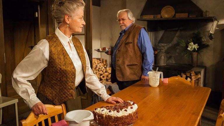 Johanna und Hermann in der Küche, auf dem Tisch steht eine Schwarzwälder Kirschtorte, Hermann hält einen Teller, mit einem Stück Torte