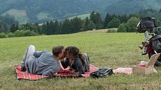 Andreas und Eva liegen auf ihrer Picknickdecke und küssen sich