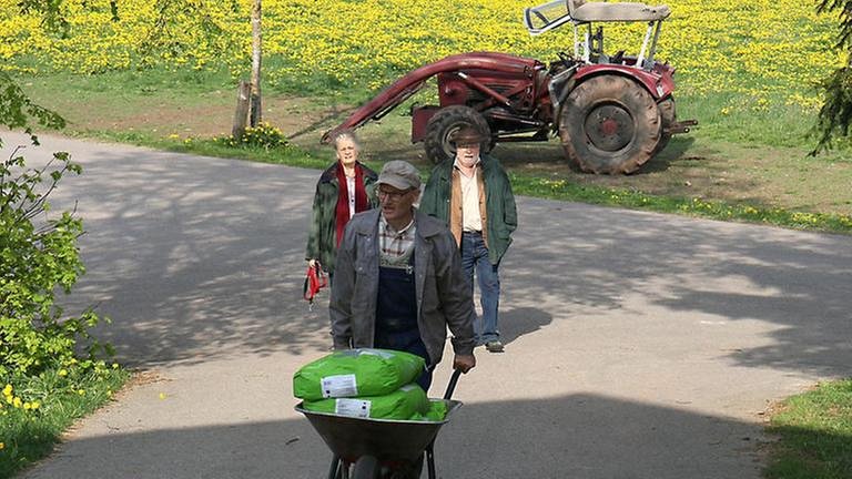 Karl schiebt die mit Säcken beladene Schubkarre, Hermann und Johanna im Hintergrund vor einem Traktor