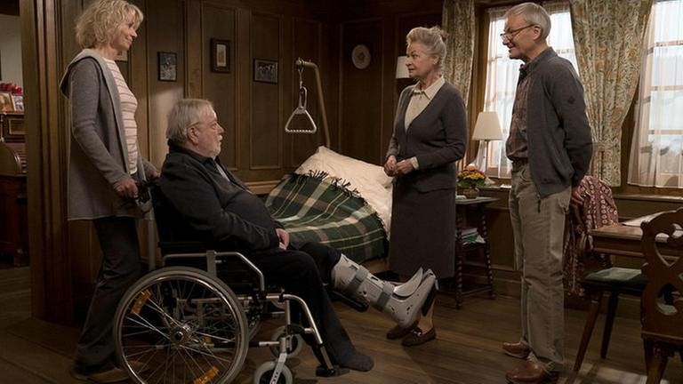Kati schiebt Hermann, der im Rollstuhl sitzt ins Wohnzimmer, wo Johanna und Karl bereits auf ihn warten