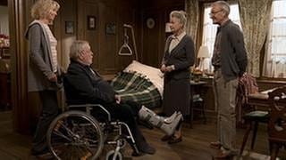 Kati schiebt Hermann, der im Rollstuhl sitzt ins Wohnzimmer, wo Johanna und Karl bereits auf ihn warten