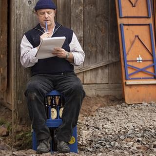 Toni sitzt mit Block und Bleistift auf einer Bierkiste vor der renovierten Mühle