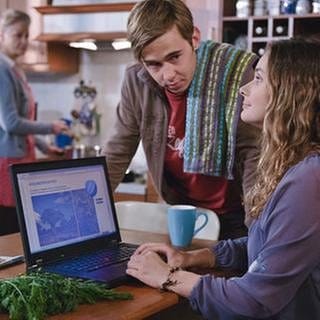 Jenny sitzt vor dem Laptop, Sebastian steht neben ihr, im Hintergrund unscharf Johanna