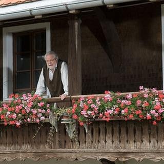 Hermann schaut vom Balkon herunter, an dem üppig die Geranien hängen