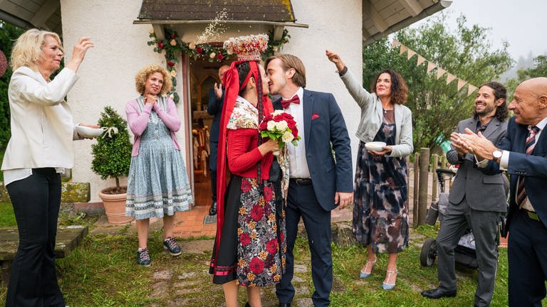 Die Hochzeitsgäste feiern das Brautpaar Jenny (Julia Obst) und Sebastian (Dominik Stricker), welches sich vor der Hofkapelle des Fallerhofes küsst.