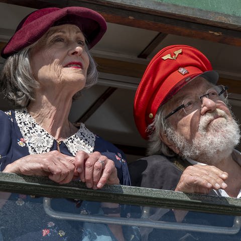 Hermann und Irene, beide mit roter Mütze und für ihren Ausflug mit der 3-Seen-Bahn schickt gemacht, stehen nebeneinander an einem geöffneten Fenster eines historischen Bahnwaggons.