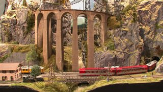 Ein gelber und ein roter Zug einer Modelleisenbahn mit Felsen und Brücke