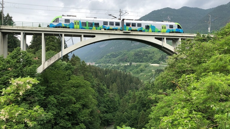 Zug der Nonstalbahn auf der Bogenbrücke über den Fluss Noce. Zu Deutsch heißt er Nonsbach oder Ulz.