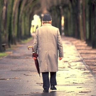 Ein älterer Herr mit einem Regenschirm läuft an einem Herbsttag alleine auf dem Gehweg.