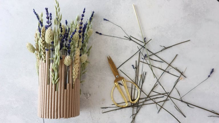 Damit die Trockenblumen nicht durch die Strohhalme rutschen, hierfür ein bisschen Heißkleber an den unteren Stiel geben und vorsichtig in den Strohhalm stecken. 