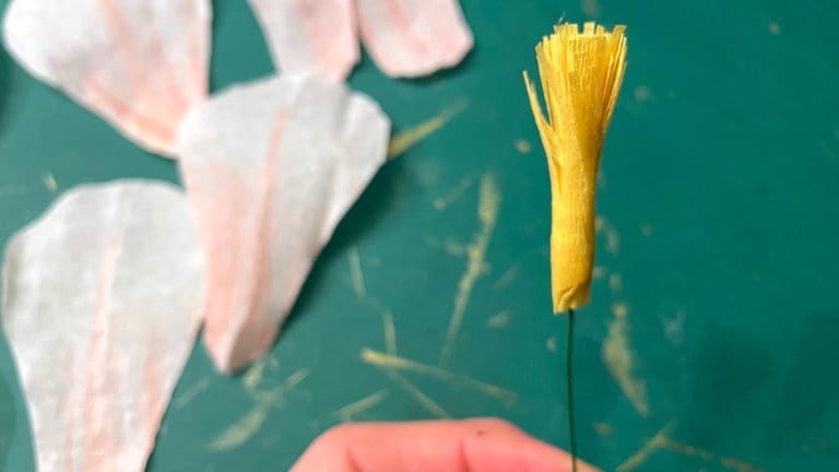 Das Ende eines Steckdrahtes oben zu einer kleinen Schlaufe biegen. Etwas Heißkeber auftragen und das gelbe Papier mit den Fransen nach oben darum wickeln. Das Ende wieder verkleben. 