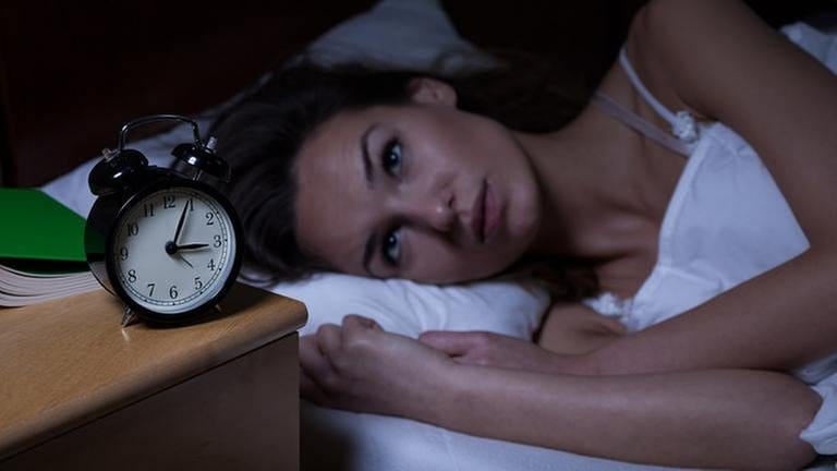Frau liegt im Bett und schaut auf ihren Wecker der vier nach drei Uhr nachts zeigt.