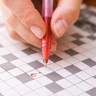 Eine Frauenhand hält einen Stift in der Hand. Sie liegt auf einem noch ungelöstem Kreuzworträtsel mit nur einem eingetragenen  Fragezeichen.