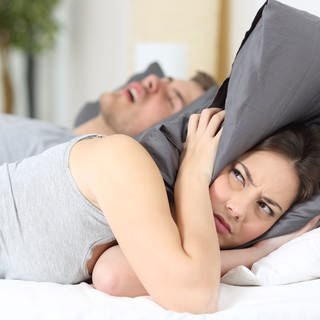 Schnarchen - Frau deckt Ohren mit Kissen zu, während Mann schläft