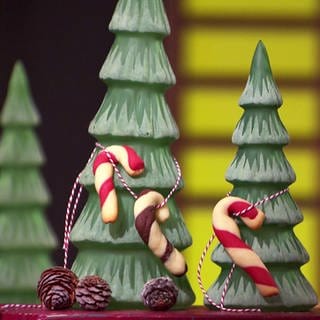 Drei Zuckerstangen-Cookies aufgehängt an zwei Deko-Tannenbäumen, im Hintergrund beleuchtete Weihnachtsmannfigur