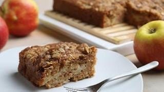 Ein leckeres Apfel-Zimt Kuchenstück liegt auf einem Teller. Im Hintergrund sieht man vereinzelt Äpfel, sowie die den Rext vom Kuchen.