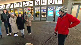Ein Mann in roter Jacke und mit Hund und eine Gruppe Menschen am Hauptbahnhof Ulm. Der ehemalige Wohnungslose Jörn "Lemmy" Zweng zeigt bei der Stadtführung "Blickwinkel" der Caritas Interessierten die Stadt Ulm aus seiner Perspektive - hier einer Gruppe von Studierenden.