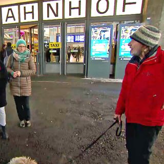 Ein Mann in roter Jacke und mit Hund und eine Gruppe Menschen am Hauptbahnhof Ulm. Der ehemalige Wohnungslose Jörn "Lemmy" Zweng zeigt bei der Stadtführung "Blickwinkel" der Caritas Interessierten die Stadt Ulm aus seiner Perspektive - hier einer Gruppe von Studierenden.