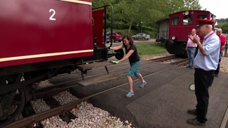 Eine Frau schiebt einen Eisenbahnwagon und ein Schaffner pfeifft 