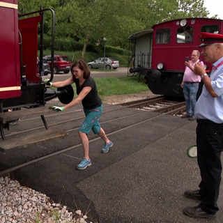 Eine Frau schiebt einen Eisenbahnwagon und ein Schaffner pfeifft 