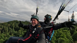 Zwei Männer sind an einem Fallschirm in der Luft und zeigen Daumen nach oben