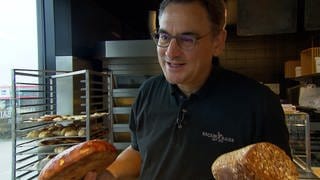 Bäcker Baier steht mit einem Brot in der Hand in seiner Bäckerei.