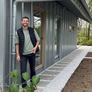 Felix steht vor seinem neuen selbst gebauten Ferienhaus und lächelt in die Kamera