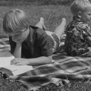 Zwei Kinder sitzen auf einer Picknickdecke, eines liest ein Buch.