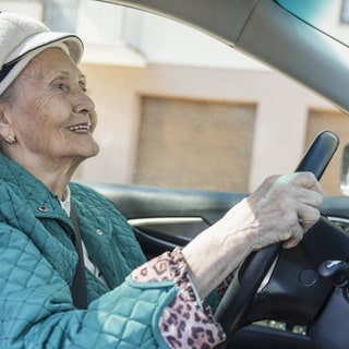 Eine Seniorin sitzt lächelnd am Steuer eines Autos.