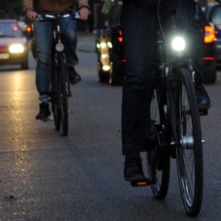 Ein Fahrradfahrer mit Licht und einer ohne sind in der Dämmerung auf einer Straße unterwegs: Welche Vorschriften müssen Fahrradleuchten erfüllen?