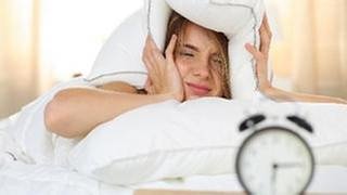 Eine Frau liegt im Bett und hält sich mit müdem Blick auf einen Wecker das Kopfkissen über die Ohren.