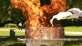 Feuerwehrmann in Schutzanzug schüttet Spiritus auf brennenden Grill mit Stichflamme. Erste Hilfe bei Verbrennungen: Soll man Brandwunden kühlen?