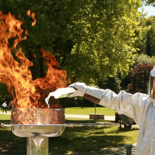Feuerwehrmann in Schutzanzug schüttet Spiritus auf brennenden Grill mit Stichflamme. Erste Hilfe bei Verbrennungen: Soll man Brandwunden kühlen?