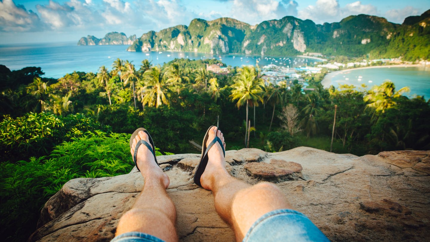 Post-Holiday-Blues: Urlauber fotostagrafit seine Beine mit Flips Flops an den Füßen auf einem Berg auf der thailändischen Insel Phi Phi. Die Aussicht kurz vor den Sonnenuntergang ist traumhaft schön.