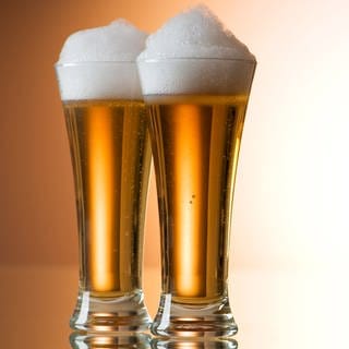 Zwei Gläser Bier stehen nebeneinander auf einem Tisch. Chemie hat einiges mit der Bildung von Bierschaum zu tun.