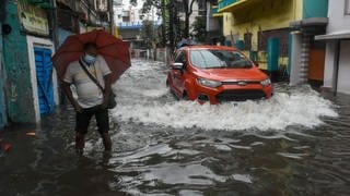COP27: Ein Auto fährt durch eine überschwemmte Straße, in der ein Mann mit Regenschirm steht. In Kalkutta ist ein Starkregen niedergegangen, der mehrere Teile der Stadt überschwemmt hat.