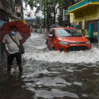 COP27: Ein Auto fährt durch eine überschwemmte Straße, in der ein Mann mit Regenschirm steht. In Kalkutta ist ein Starkregen niedergegangen, der mehrere Teile der Stadt überschwemmt hat.
