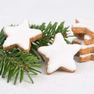 Der Weihnachtsklassiker Zimtsterne lassen sich schnell und einfach aus einer Marzipanrohmasse zubereiten. Sechs Zimtsterne liegen auf einem Tannenzweig.