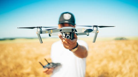 Mann mit weißem T-Shirt und Baseball-Cap hält Drohne in die Kamera. Wie viel sollte eine vernünftige Drohne kosten und wer darf sie fliegen?